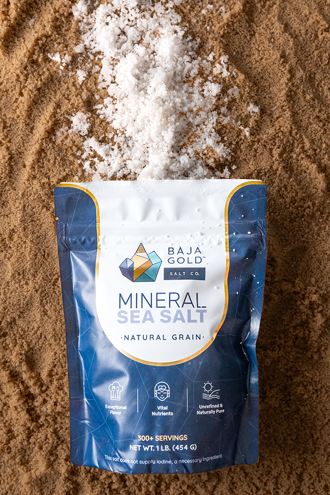 Natural grain Baja Gold mineral sea salt package spilling onto a sandy background.